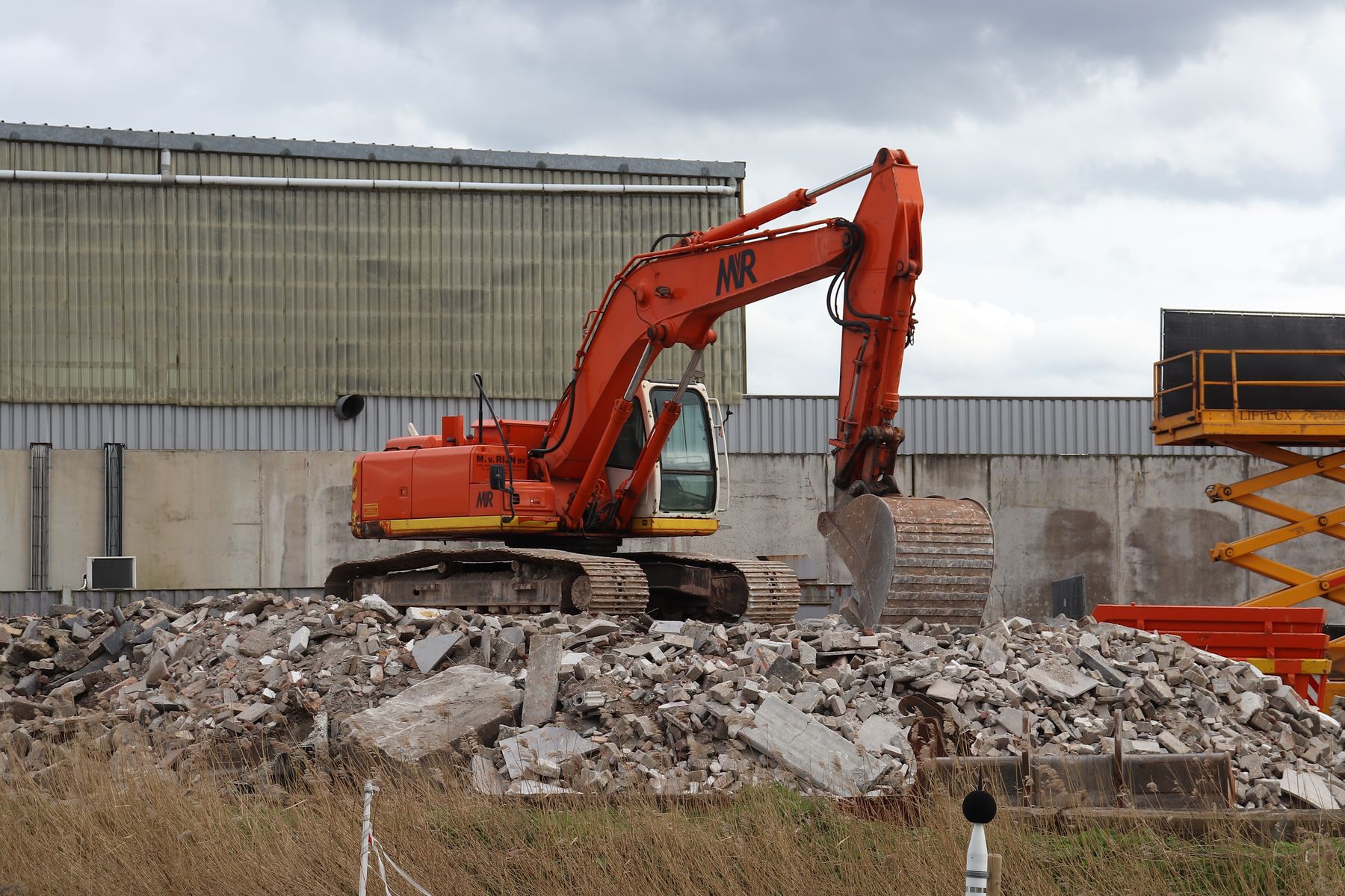 An orange excavator sitting on a pile of broken concrete beside a metal-clad warehouse.
